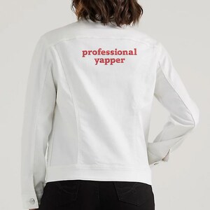 May include: White denim jacket with red embroidered text on the back that reads "professional yapper"