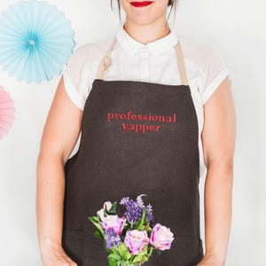 May include: A brown linen apron with a white adjustable strap and the text "professional yapper" embroidered in red. The apron has a large pocket on the front and is being worn by a person holding a bouquet of pink and purple flowers.