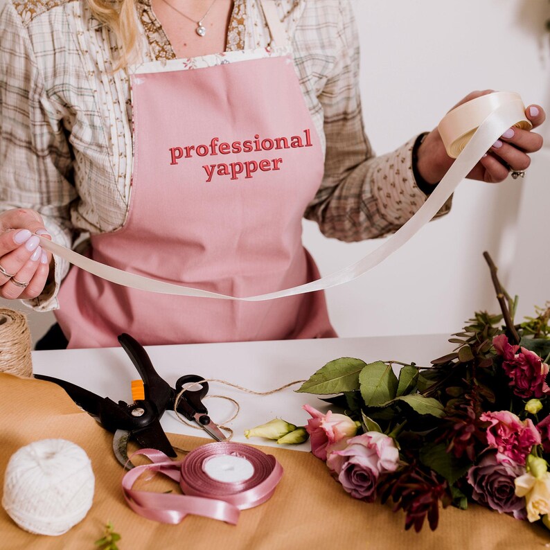 May include: A person wearing a pink apron with the text "professional yapper" embroidered on it, holding a white ribbon. The person is working on a floral arrangement with pink, purple, and white flowers.