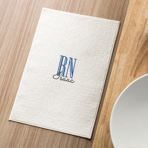 May include: White placemat with blue embroidered initials "RN" and the name "Isaac" underneath.