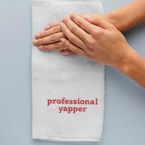 May include: White hand towel with the words "professional yapper" embroidered in red.