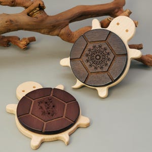 May include: Two wooden turtle-shaped coasters. The coasters have a light wood base with a dark wood, hexagonal top. One coaster features a floral design, and the other has a mandala design. The coasters are on a gray surface.