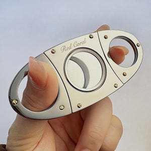 Può includere: Un taglia sigari in argento con finiture dorate e le parole "Red Coral" incise sulla parte superiore. Il taglia sigari ha due lame e un foro rotondo per tagliare i sigari.