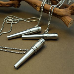 May include: Close-up of three silver-colored metal pendant necklaces. Each pendant has a cylindrical design with textured detailing on the ends. The necklaces are displayed on a brown surface with a wooden branch in the background.