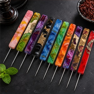 May include: A collection of ten colorful, decorative skewers with long, thin metal needles. The handles are rectangular and feature a variety of vibrant colors and patterns, including pink, green, purple, black, blue, orange, and red. The skewers are arranged on a dark surface.