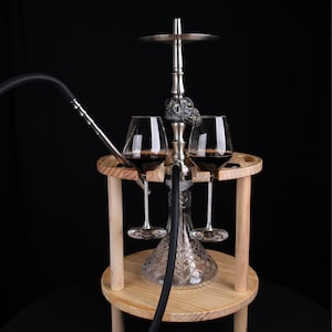 May include: A silver hookah with a glass base and a black hose, set on a wooden stand. Two wine glasses filled with red liquid are placed on the stand. The hookah has decorative skull accents.