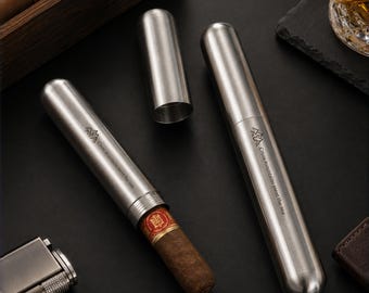 May include: Two cylindrical stainless steel cigar cases with a brushed metal finish. One case is closed, while the other is open, revealing a brown cigar inside. Both cases feature a small mountain icon and the text Cross mountains meet the sea.