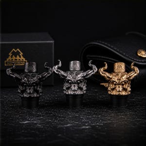 May include: Three decorative bottle stoppers with a horned skull design. The stoppers are in black, silver, and gold colors. The background includes a black box with a mountain logo and a black leather wallet.