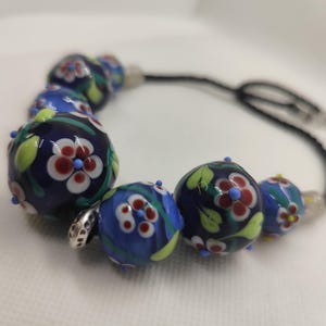 Op de afbeelding: Een handgemaakte armband met donkerblauwe, ronde glazen kralen. Elke kraal is versierd met een bloemmotief in rood, wit en groen. De armband is geregen op een zwart koord en heeft een zilverkleurige sluiting. De kralen zijn ongeveer 2 cm in diameter.
