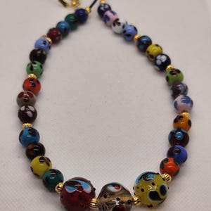 Può includere: Una colorata collana di perline con una varietà di perline rotonde decorate nei toni del rosso, blu, giallo, verde e bianco. Le perline sono infilate su un cordino nero con dettagli dorati.
