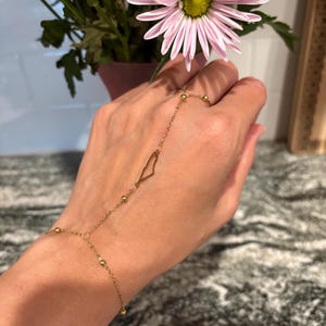 Puede incluir: Una delicada pulsera de cadena de mano dorada. La pulsera presenta una cadena que se conecta a un anillo y una cadena que se conecta a la muñeca. El diseño incluye pequeñas cuentas doradas y una forma geométrica en la mano.