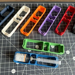 May include: A set of seven 3D printed plastic organizers in various colors, including blue, white, orange, purple, red, green, and black. The organizers have three compartments labeled with numbers. A clear glass vial with a needle is inserted into the blue organizer.