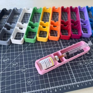 May include: A set of 10 3D printed storage organizers in various colors, including black, gray, white, green, yellow, orange, red, pink, purple, and blue. The organizers have three compartments and are designed to hold small items, such as insulin pens or other medical supplies. A pink organizer is shown with a syringe and a vial of insulin.