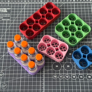 May include: Five different 3D printed bottle organizers in various colors, including red, green, pink, and blue. The organizers are made of plastic and have a honeycomb design. The organizers are sitting on a black cutting mat with white markings.