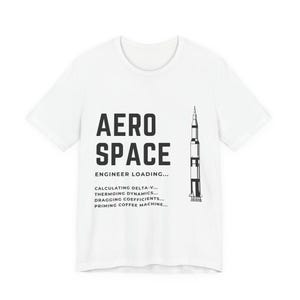 May include: A white short-sleeve t-shirt featuring a black graphic of a rocket and the text AERO SPACE ENGINEER LOADING... CALCULATING DELTA-V... THERMOING DYNAMICS... DRAGGING COEFFICIENTS... PRIMING COFFEE MACHINE... printed on the front chest area.