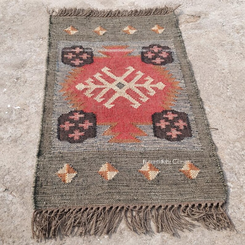 May include: A rectangular jute rug with a central geometric design in red, beige, and orange. The rug has a gray border with diamond and floral patterns, and fringe along the bottom edge. The rug measures approximately 24 x 36 inches.