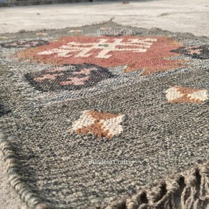 May include: A gray and brown woven rug with a geometric pattern. The rug features a central design in red and white, surrounded by dark gray and brown accents. The edges have a fringe detail. The text "Bundela Crafts" is visible.