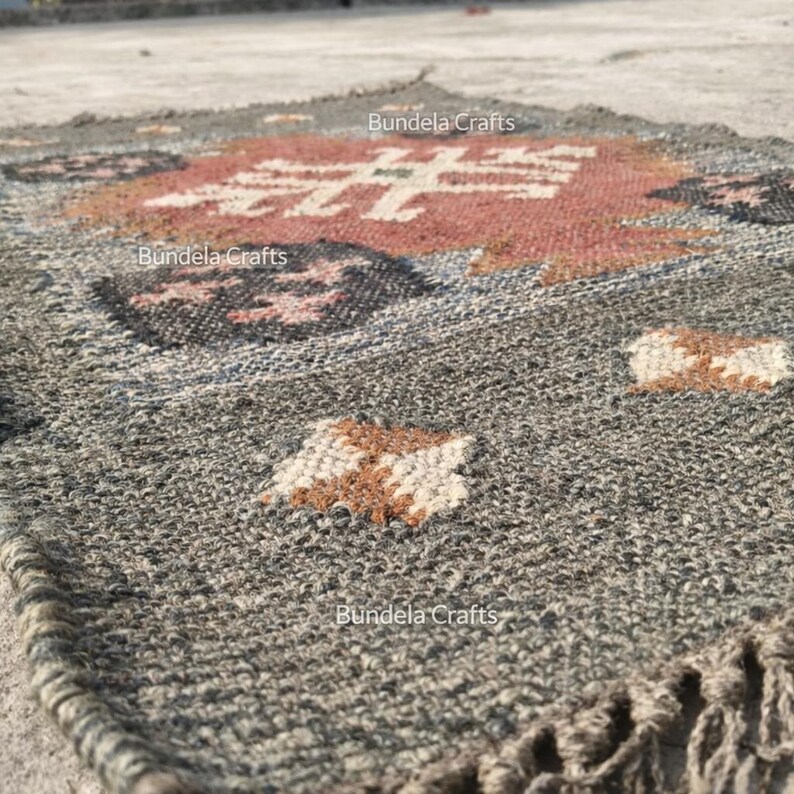 May include: A gray and brown woven rug with a geometric pattern. The rug features a central design in red and white, surrounded by dark gray and brown accents. The edges have a fringe detail. The text "Bundela Crafts" is visible.