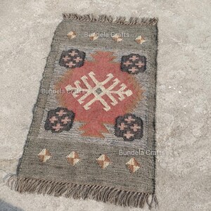 May include: A rectangular gray rug with a geometric design in red, black, and beige. The rug has a fringed edge and the text "Bundela Crafts" is visible. The rug is likely made of natural fibers.
