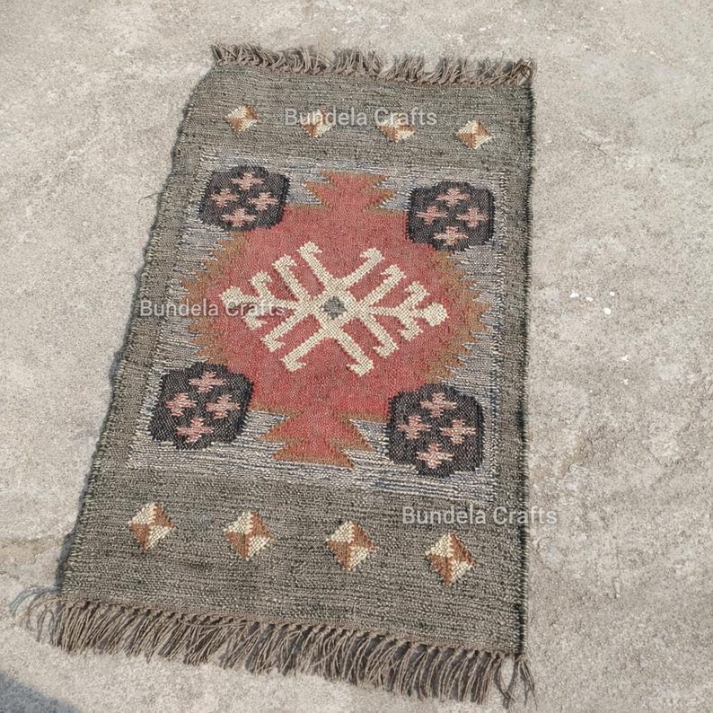 May include: A rectangular gray rug with a geometric design in red, black, and beige. The rug has a fringed edge and the text "Bundela Crafts" is visible. The rug is likely made of natural fibers.
