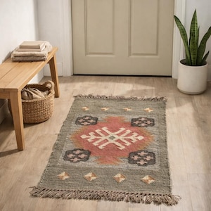 May include: A rectangular, olive green rug with a central geometric design in coral, cream, and brown. The rug has a fringed edge and is placed on a light wood floor. The rug is a decorative floor covering.