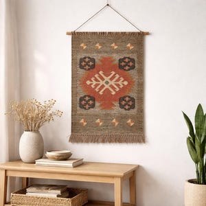May include: A rectangular wall hanging with a brown background and a central geometric design in orange, cream, and black. The tapestry is suspended from a wooden rod and has a fringed bottom. The wall decor is displayed on a white wall.