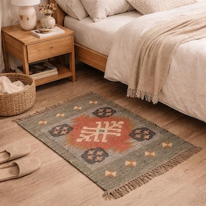May include: A rectangular area rug with a geometric design in shades of green, orange, and gray. The rug has a fringed edge and is placed on a wooden floor near a bed and nightstand.