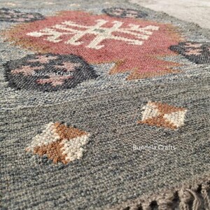 May include: A gray woven rug with a geometric pattern in shades of red, brown, and white. The rug features a central design and repeating motifs. The text "Bundela Crafts" is visible on the rug.