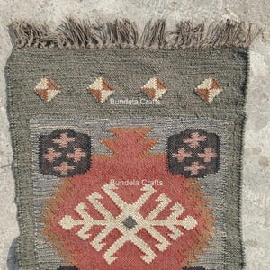 May include: A rectangular, hand-woven rug with a geometric design. The rug features a central pattern in shades of red, beige, and brown, framed by a gray border. The top edge has a fringe. The text "Bundela Crafts" is visible.