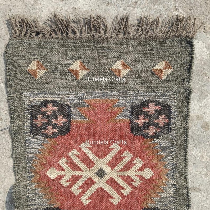 May include: A rectangular, hand-woven rug with a geometric design. The rug features a central pattern in shades of red, beige, and brown, framed by a gray border. The top edge has a fringe. The text "Bundela Crafts" is visible.