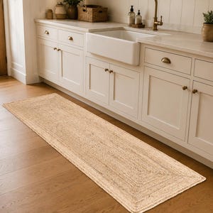 May include: A rectangular, natural-toned jute rug lies on a hardwood floor in front of a white kitchen cabinet. The rug has a woven texture and a bordered design. The kitchen features a farmhouse sink and brass hardware.