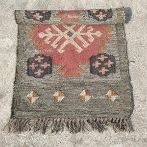 May include: A rectangular, hand-woven rug with a geometric design. The rug features a central red and beige pattern, framed by a gray border with diamond and floral motifs. The rug has a fringed edge and the text "Bundela Crafts".