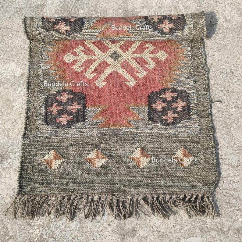 May include: A rectangular, hand-woven rug with a geometric design. The rug features a central red and beige pattern, framed by a gray border with diamond and floral motifs. The rug has a fringed edge and the text "Bundela Crafts".