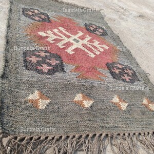 May include: A rectangular gray jute rug with a central geometric design in red, white, and brown. The rug features a fringe along the bottom edge and decorative patterns. The words "Bundela Crafts" are printed on the rug.
