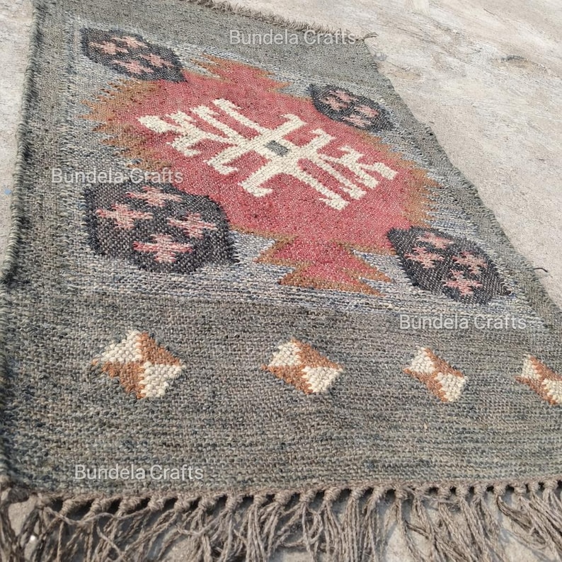 May include: A rectangular gray jute rug with a central geometric design in red, white, and brown. The rug features a fringe along the bottom edge and decorative patterns. The words "Bundela Crafts" are printed on the rug.