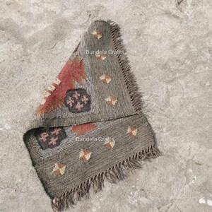May include: A folded, rectangular rug with a gray and red geometric pattern. The rug has a fringed edge and the text "Bundela Crafts" is visible. The rug is made of a woven material and is lying on a concrete surface.