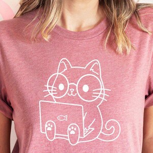 May include: A dusty rose t-shirt featuring a white outline graphic of a cat wearing glasses and using a laptop. The cat is sitting with its paws on the laptop. The background includes pink heart decorations.