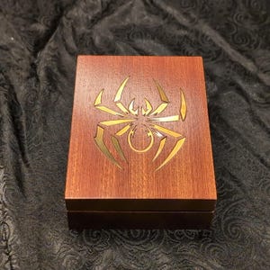 May include: A wooden box with a hinged lid. The box is a reddish-brown color and features a gold-colored spider design on the top. The spider design is intricately detailed and adds a decorative element to the box.