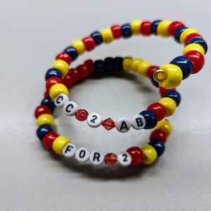May include: Two beaded bracelets with red, yellow, and blue beads. The bracelets spell out "FOR 2" and "C 2 AB".