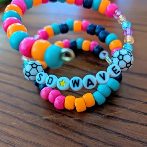 Peut inclure: Deux bracelets en perles colorées avec les mots "SD WAVE" écrits en lettres blanches avec une étoile jaune entre les mots. Les bracelets sont faits de perles turquoise, roses, orange et noires.