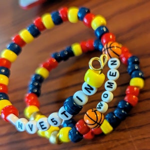 May include: Two beaded bracelets with a basketball charm and the words "Invest in Women" and "Win" spelled out in white beads.