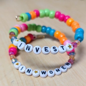 May include: A colorful beaded bracelet with the words "INVEST IN WOMEN" spelled out in white letters with gold stars between each letter.