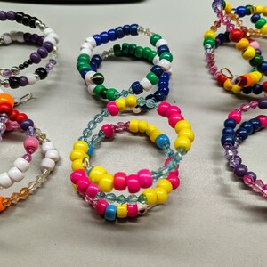 May include: Six colorful beaded bracelets, each with a different color scheme and pattern. The bracelets are made with small, round beads in various colors, including purple, green, yellow, blue, orange, and white.