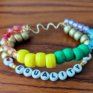 May include: A colourful beaded bracelet with a gold wire base. The beads are in a rainbow pattern and spell out "EQUALITY" with white letter beads. There are also two small red heart beads.