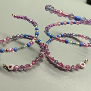 May include: Two beaded bracelets with pink, blue, and white beads. Each bracelet has a small white charm with a red heart on it.