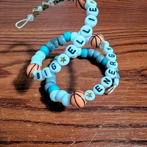 May include: Three beaded bracelets with white, gray, and turquoise beads. The bracelets feature orange basketball-shaped beads and letter beads spelling out "ELLIE" and "BIG ENERGY". Gold star-shaped beads are also included.