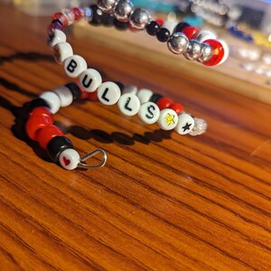May include: A beaded bracelet with the word "BULLS" spelled out in white letter beads. The bracelet features red, black, silver, and clear beads, with a star charm and a heart-shaped bead. The bracelet is on a wooden surface.