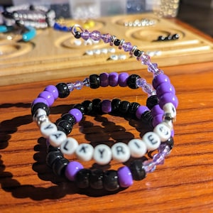 May include: Two beaded bracelets with black and purple beads. The top bracelet has the word "VALKYRIE" spelled out in white beads.