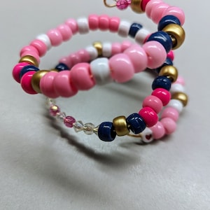 May include: Three beaded bracelets in pink, white, navy blue, and gold. The bracelets are stacked on top of each other and are coiled into a spiral shape.