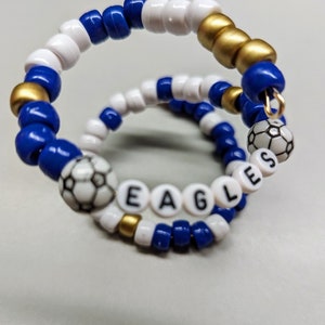 Peut inclure: Un bracelet en perles bleues, blanches et dorées avec le mot "EAGLES" écrit en perles blanches et deux breloques de ballon de football.